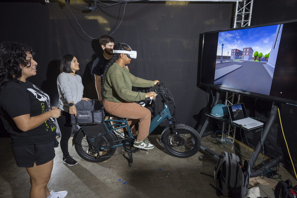 A college student with a virtual reality headset on sits on a stationary bicycle in front of a screen that shows the road experience she is engaged with.