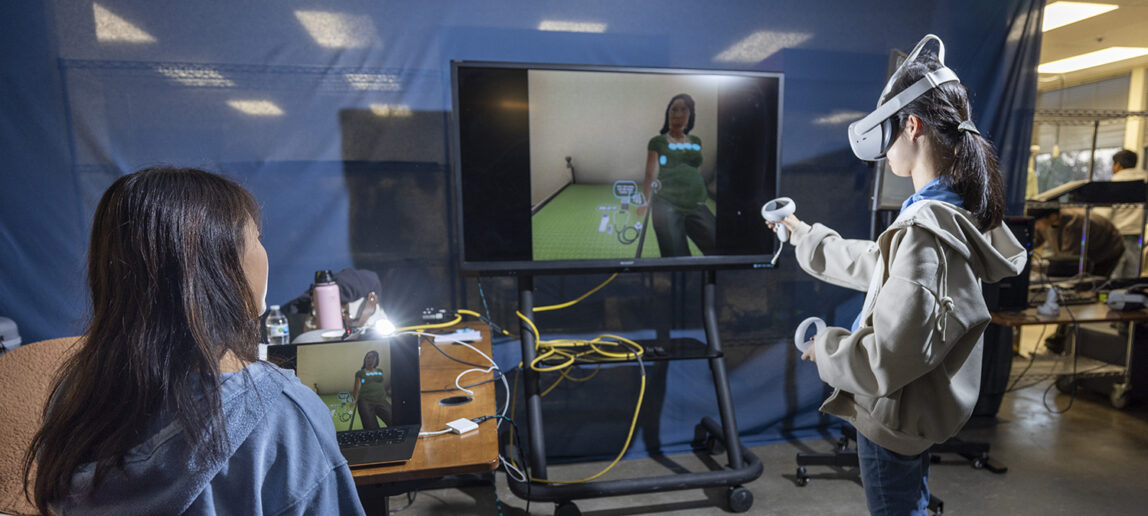 A young woman with a virtual reality headset on uses two controllers in front of a screen.