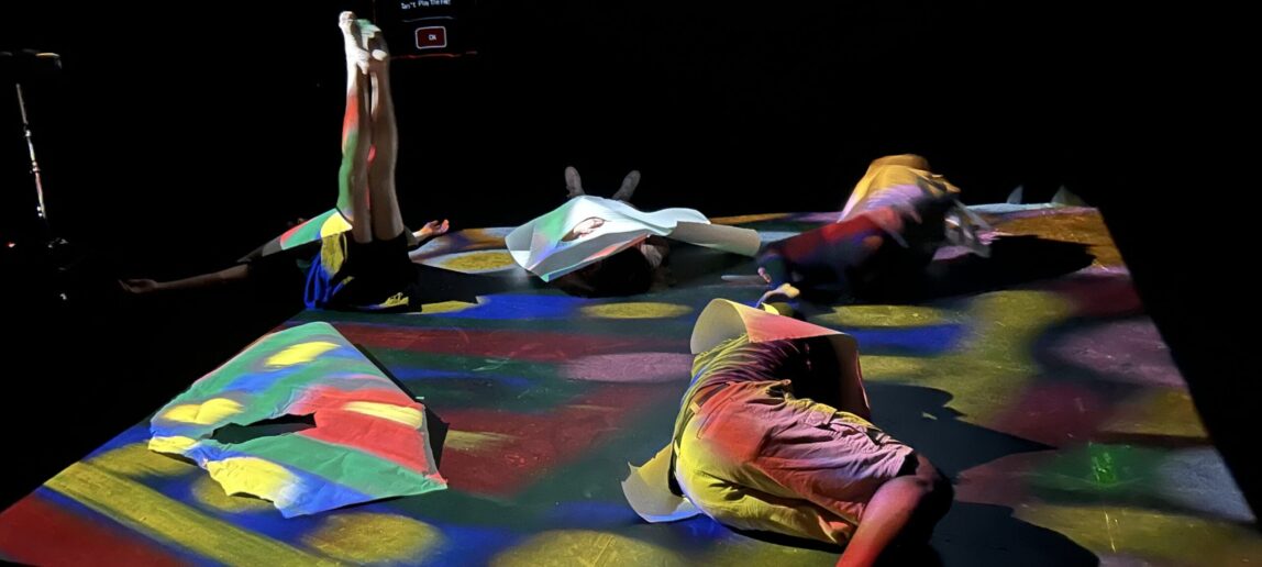 Four people lay in varying positions on the floor, which is lit in multicolored shapes from above.