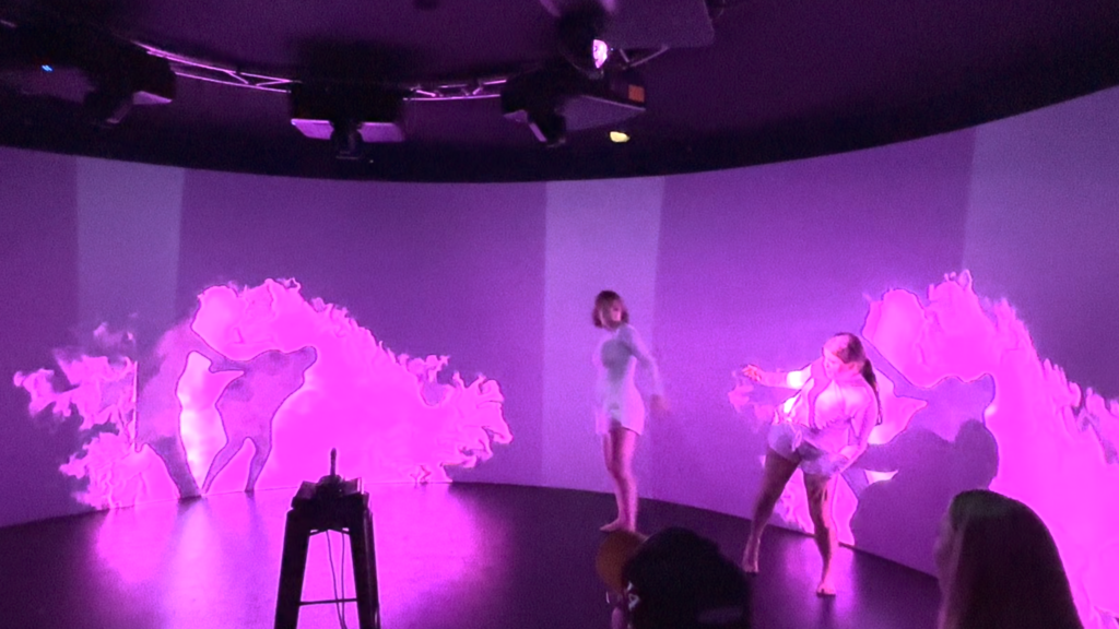 Under purple light, two dancers perform in front of a 360-degree screen while projections are displayed behind them.