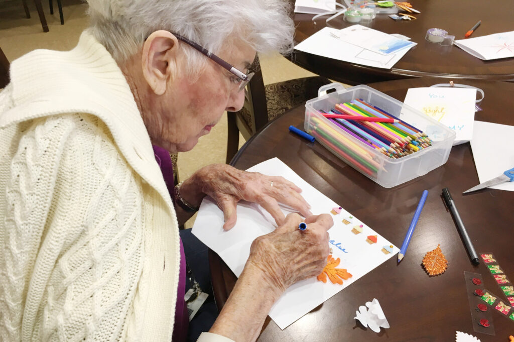 A woman sits at a table with craft supplies, writing and decorating a paper card.