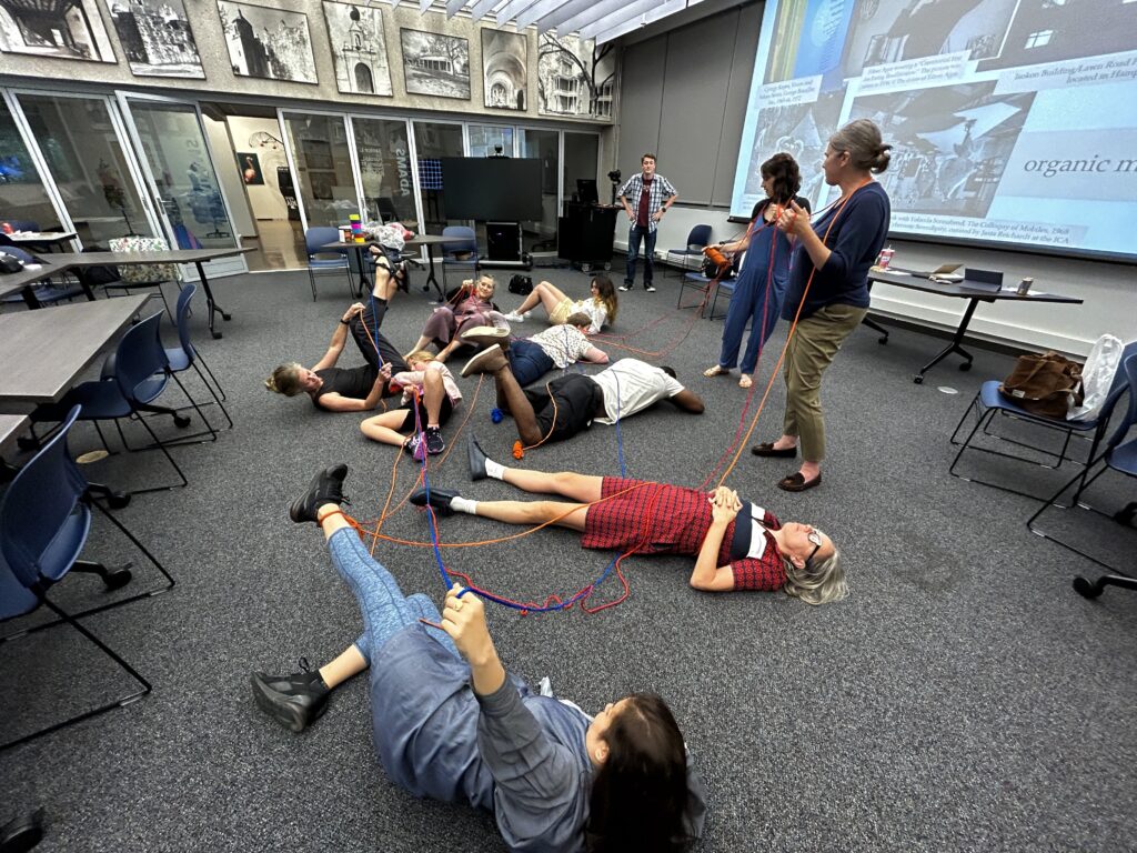 Seven people lay on the floor, each holding long strands of string, while three people stand near them in an art workshop.