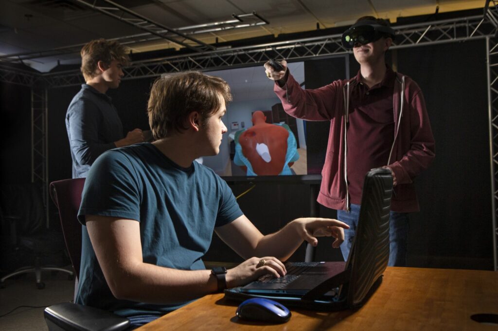 A college student wears a virtual reality headset and gestures with a hand controller. A student is seated, pointing toward a computer screen. Another student is standing. A screen displaying digital imagery is in the background.