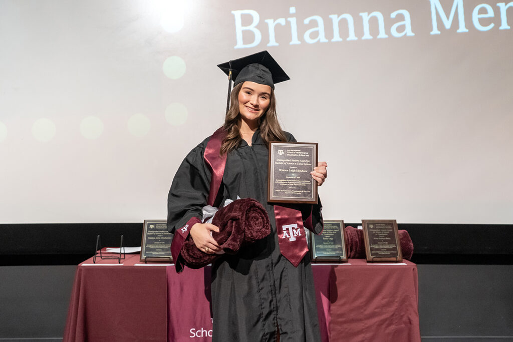 A graduating college student in a traditional cap and gown and a maroon Texas A&M sash holds a plaque on a stage.