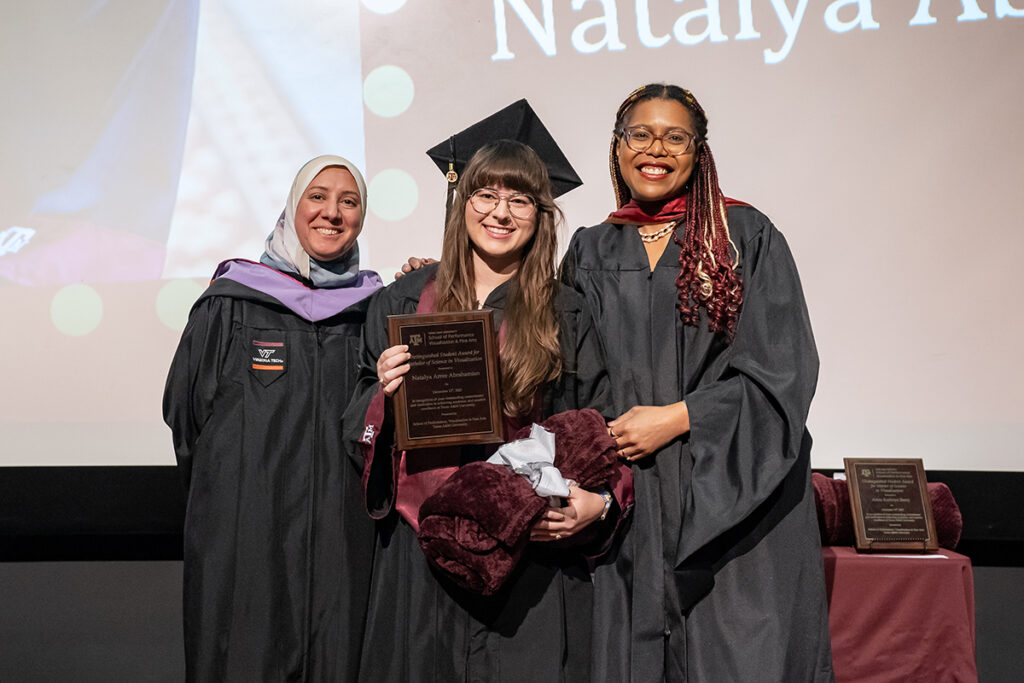A graduating college student in a traditional cap and gown and a maroon Texas A&M sash holds a plaque on a stage. On either side of her are university professors.