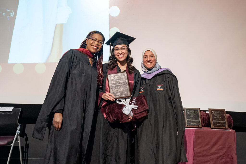 A graduating college student in a traditional cap and gown and a maroon Texas A&M sash holds a plaque on a stage. On either side of her are university professors.