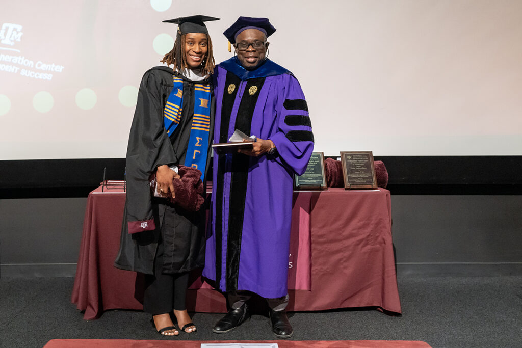 A graduating college student in a traditional cap and gown and a blue and gold sash stands on a stage. On her left is a university professor holding a plaque.