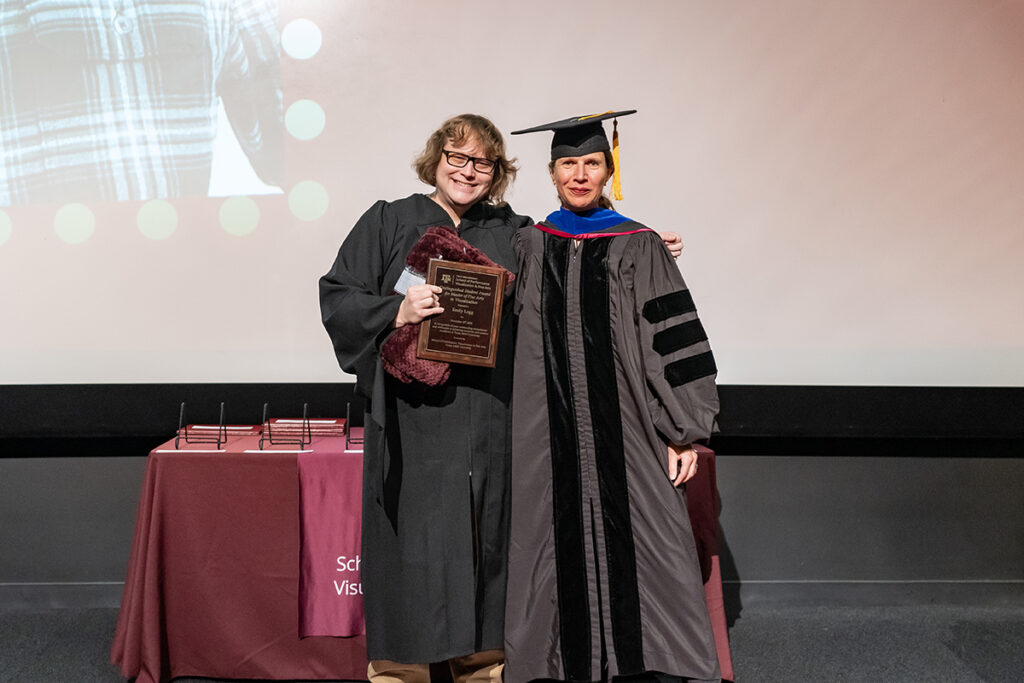 A graduating college student in a traditional cap and gown holds a plaque on a stage. On the student's left is a university professor in a cap and gown.