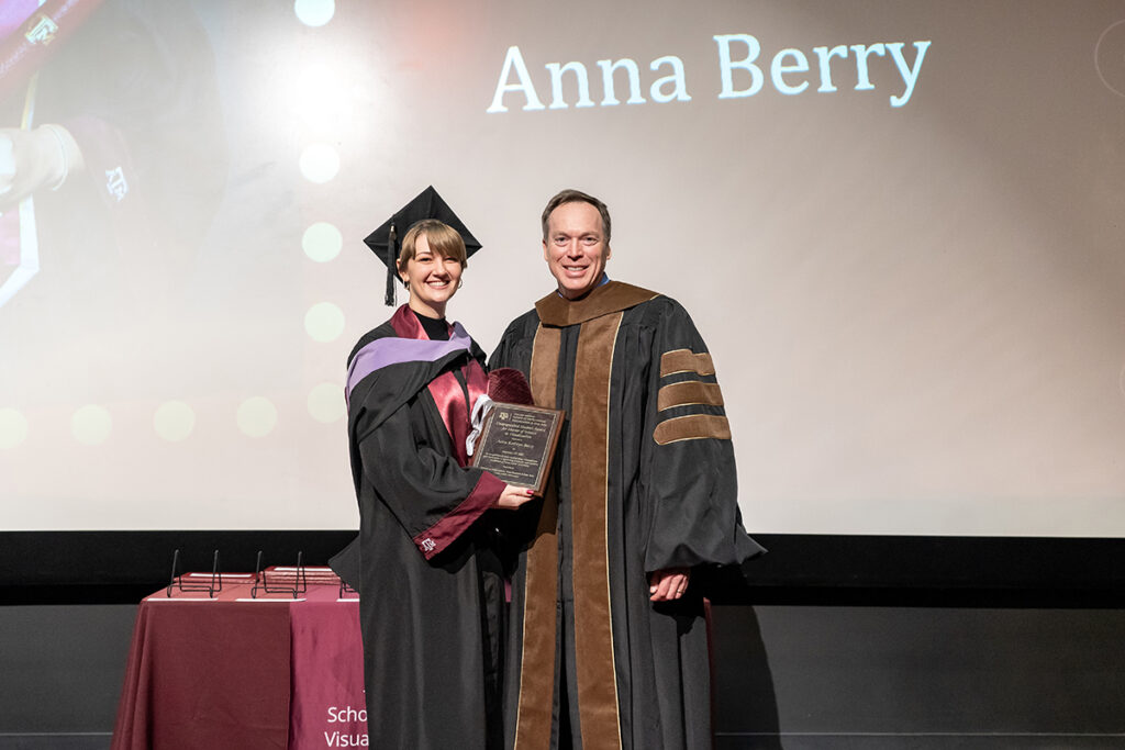 A graduating college student in a traditional cap and gown and a maroon Texas A&M sash and a purple sash holds a plaque on a stage. On her left is a university dean.