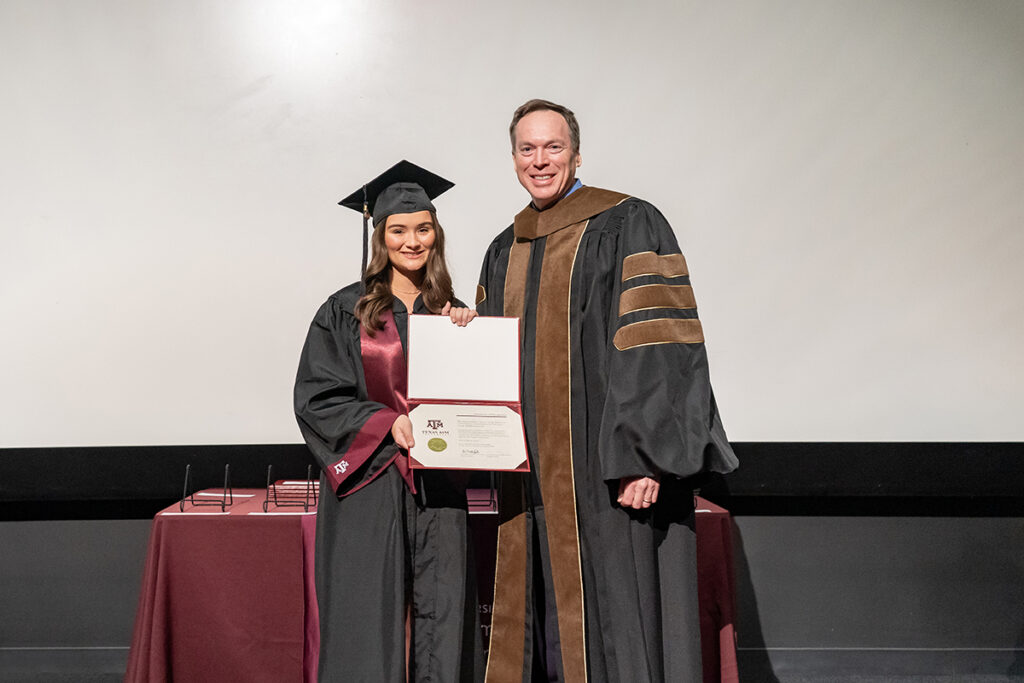 A graduating college student in a traditional cap and gown and a maroon Texas A&M sash holds a certificate on a stage. On her left is a university dean.