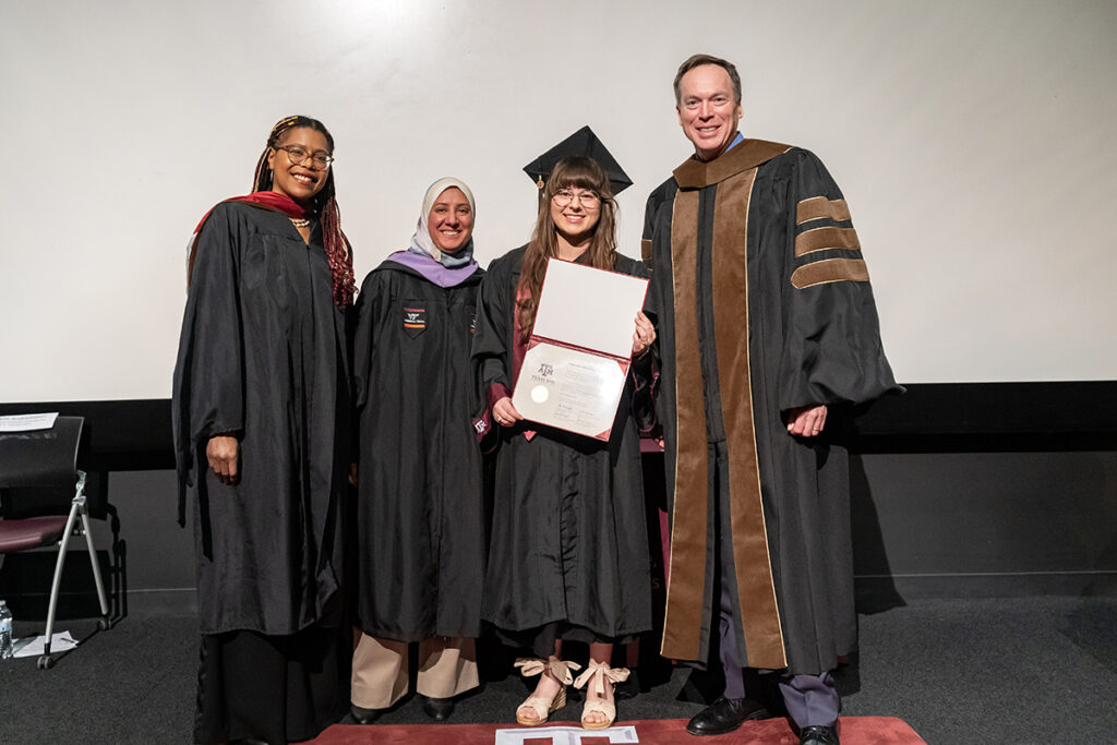 A graduating college student in a traditional cap and gown and a maroon Texas A&M sash and a purple sash holds a plaque on a stage. On her left is a university dean, and on her right are two university professors, all wearing caps and gowns.