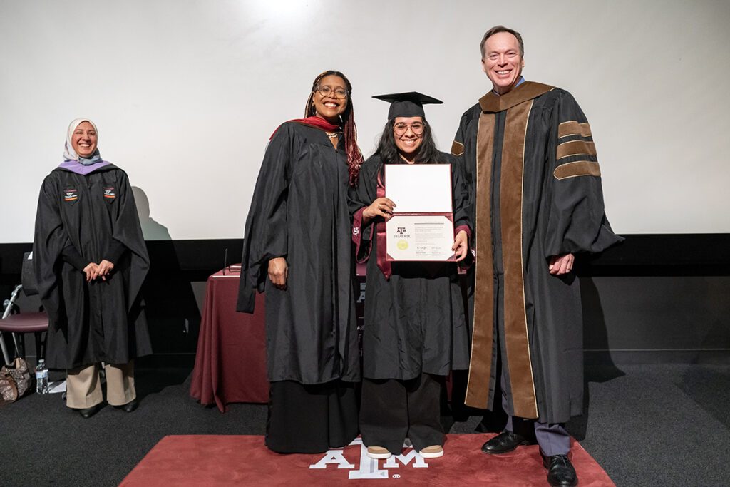 A graduating college student in a traditional cap and gown and a maroon Texas A&M sash holds a plaque on a stage. On her left is a university dean and on her right is a university professor, all wearing caps.