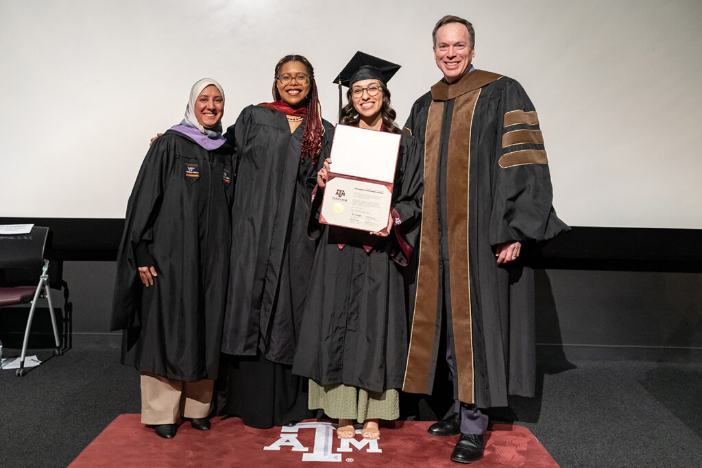 A graduating college student in a traditional cap and gown holds a certificate on a stage. On her left is a university dean, and on her right are two university professors, all wearing graduation gowns.