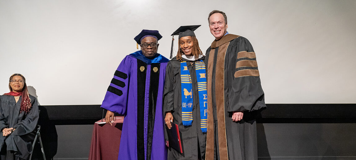 A graduating college student in a traditional cap and gown and a blue and gold sash holds a plaque on a stage. On her left is a university dean, and on her right is a university professor, all wearing graduation gowns.
