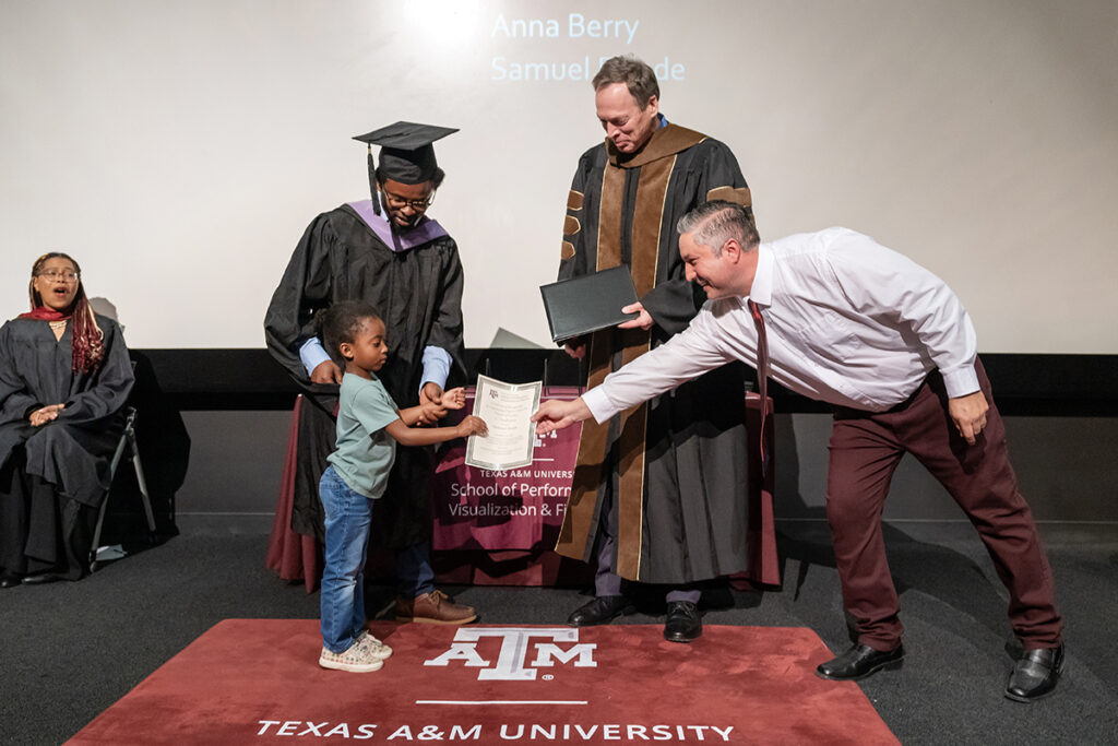 A graduating college student in a traditional cap and gown looks down at his child, who is being given a certificate by a man in a tie on the right. A university dean smiles as he looks at the child.