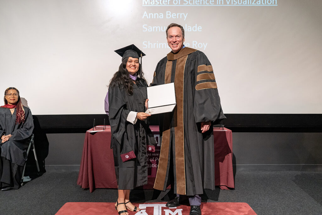 A graduating college student in a traditional cap and gown and a purple sash holds a certificate on a stage. On her left is a university dean.