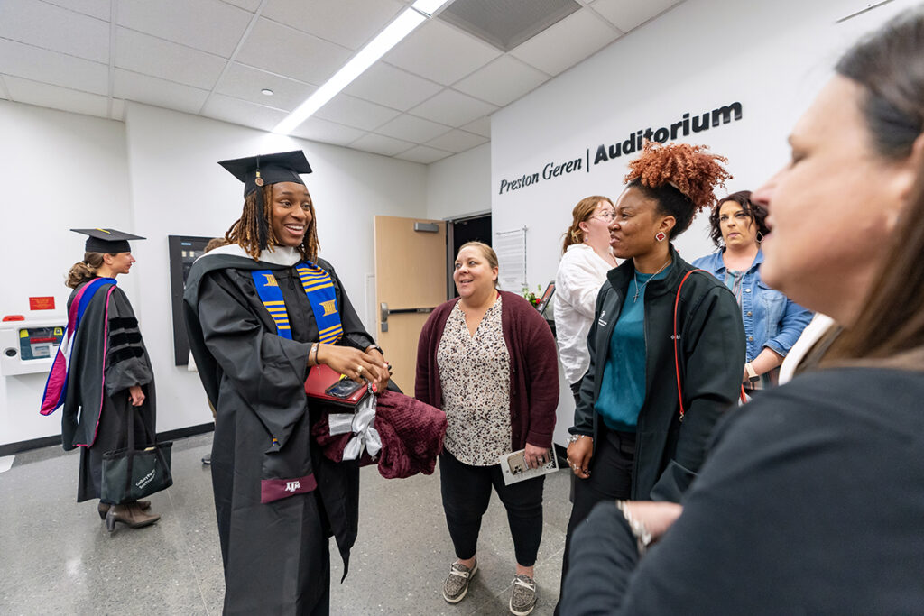 A graduating college student in a traditional cap and gown and a blue and yellow sash smiles as she speaks with others after a graduation ceremony.