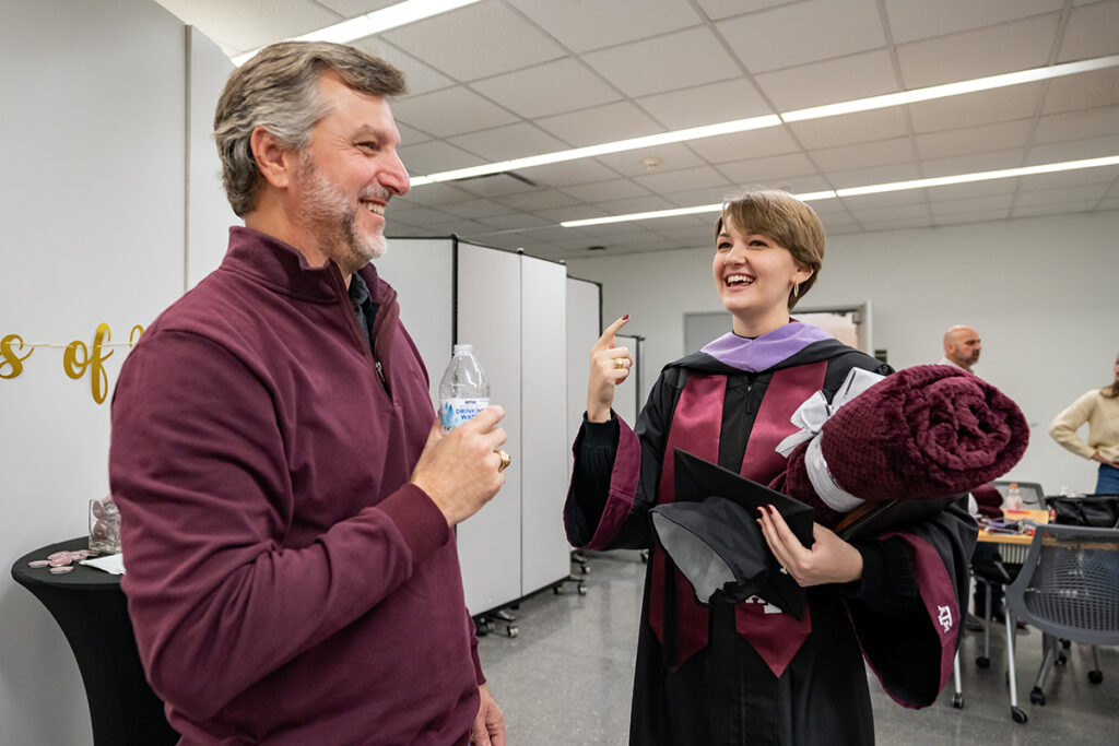 A graduating college student in a traditional graduation gown and a maroon Texas A&M sash and a purple sash smiles as she speaks with a man on her right after a graduation ceremony.