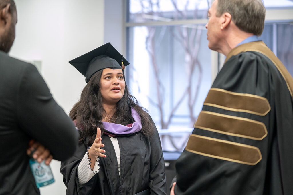 A graduating college student in a traditional cap and gown and a purple sash speaks with a university dean after a graduation ceremony.