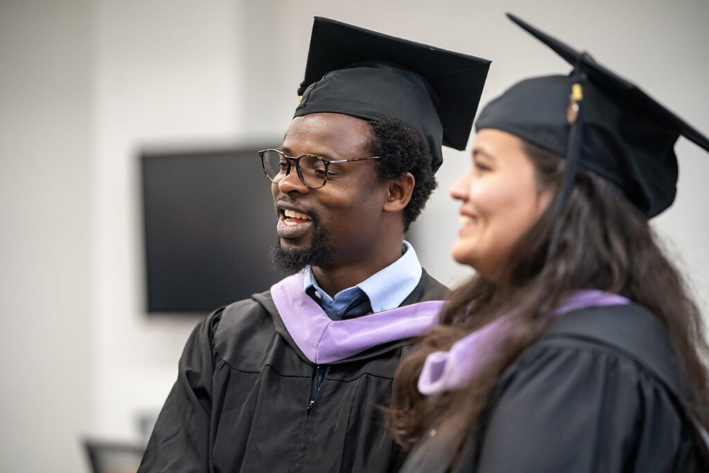 Two graduating college students in traditional caps and gowns and purple sashes smile as they talk after a graduation ceremony.