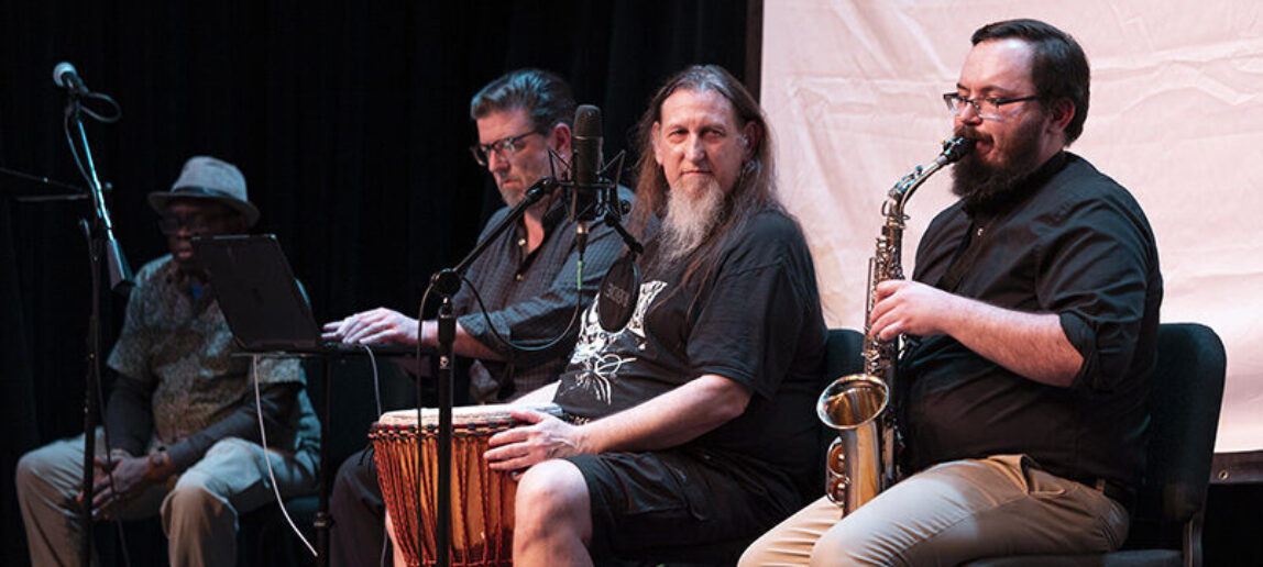 Four men are sitting on a stage. Furthest left is a college professor wearing a hat. Second from left is a college professor working with a computer. Third from left is a college professor with bongo drums. At right is a college staff member playing a saxophone.