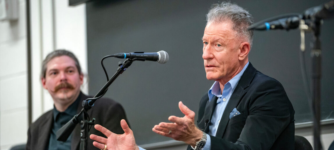 Singer-songwriter Lyle Lovett is shown speaking to college students in a classroom. Lovett is seated and a microphone is set up in front of him. To his right is the class professor, Matthew Campbell.