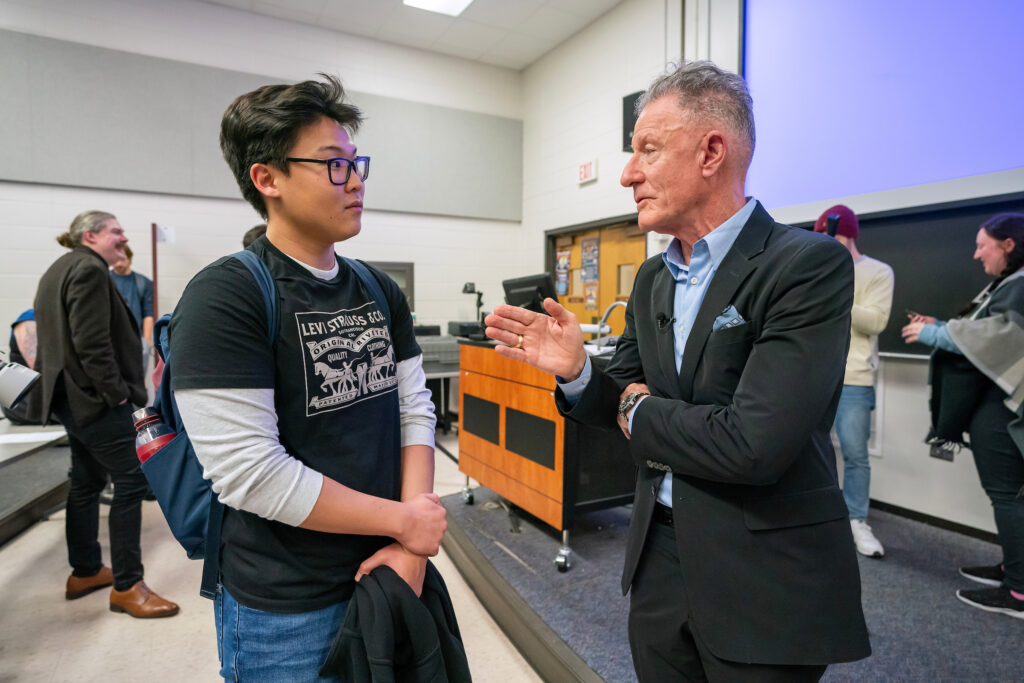 Singer-songwriter Lyle Lovett speaks to a college student in a classroom. Students and a professor are behind them.