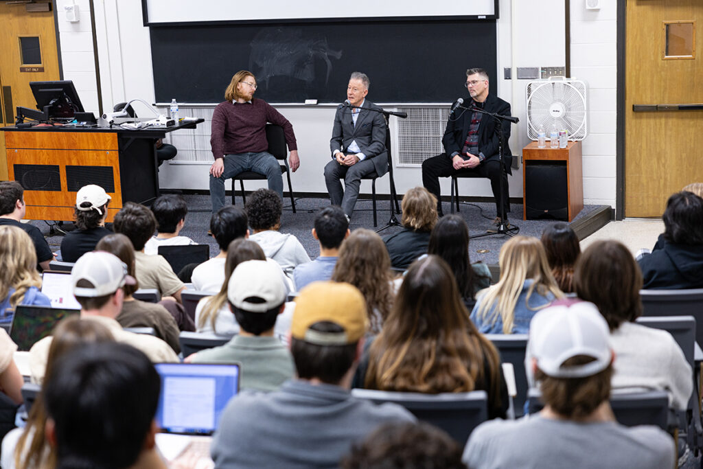 Lyle Lovett sits on a stage in front of a college class, speaking to students. on his right is the class professor. On his left is his tour manager. 