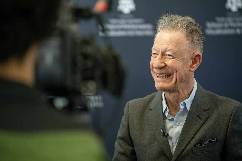 Singer-songwriter Lyle Lovett speaks to local media, in front of a backdrop that has the Texas A&M School of Performance, Visualization and Fine Arts logo.