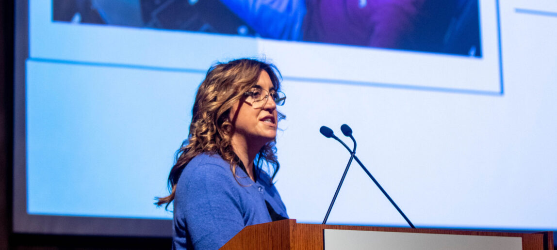 A university professor wearing a blue sweater stands at a podium that is labeled Texas A&M Annenberg Presidential Conference Center. The Bush School. Texas A&M University.