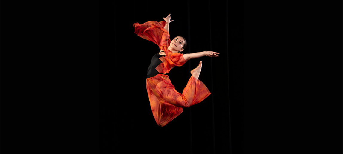 A dancer wearing an orange and black outfit is seen midair, with her legs extended beyond her back, and her arms reaching outward.