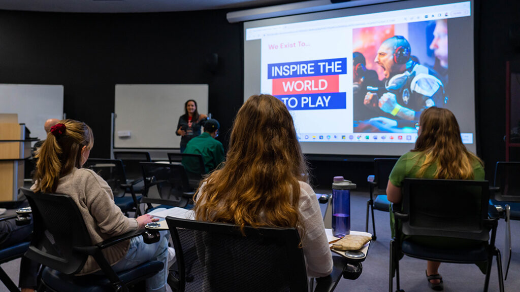 Four college students sit at desks and view a presentation led by a video game professional. On the screen is a gaming scene with the words "We Exist To ... Inspire the World to Play."