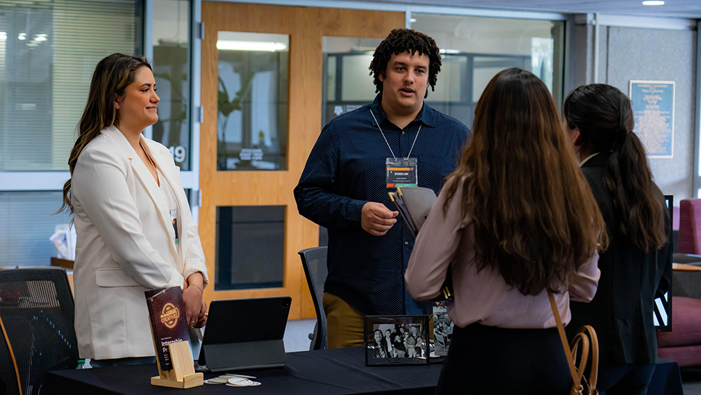 Two professionals standing at a resource table talk with two college students at a job fair.
