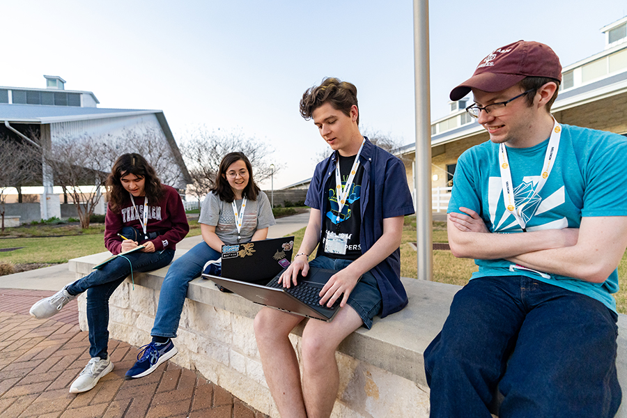 Four college students sit outside and discuss a video game project. Two of the students are working on laptop computers.