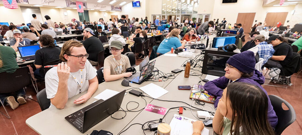 Four college students seated at a table smile and talk at a video game jam.