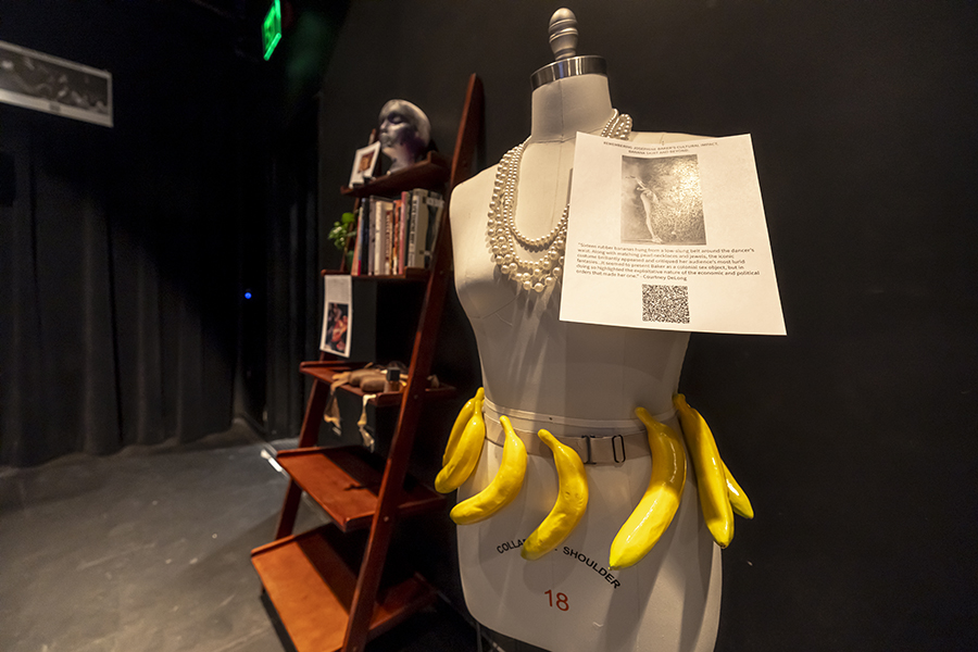 A bookshelf with Black dance history books is shown. Next to it is a mannequin body, with an elaborate necklace and a belt with banana shapes.