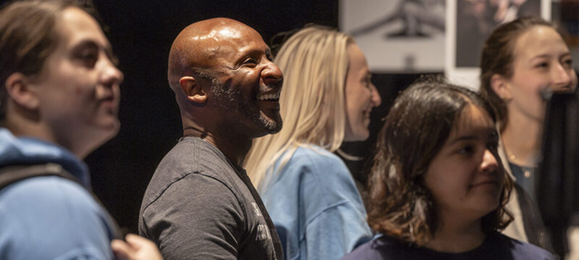 A college dance professor surrounded by students smiles as he watches a dance display at an interactive exhibit.