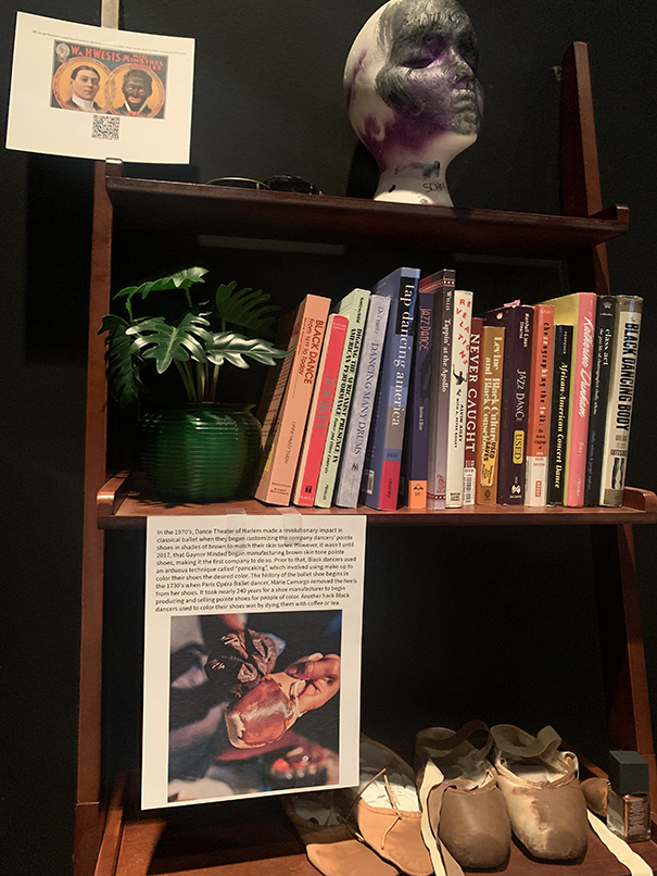 A bookshelf on display in an art exhibit is shown, including books and ballet shoes that have been painted.