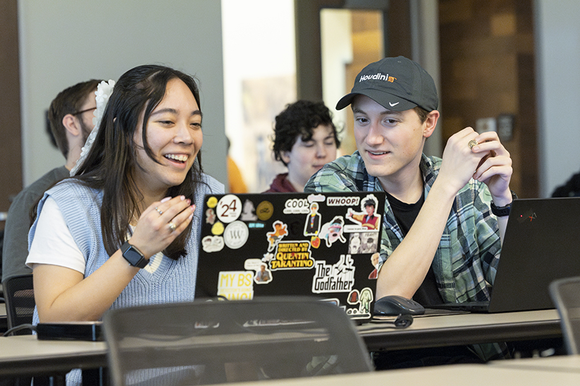 Two college students smile as they talk while looking at a computer screen.