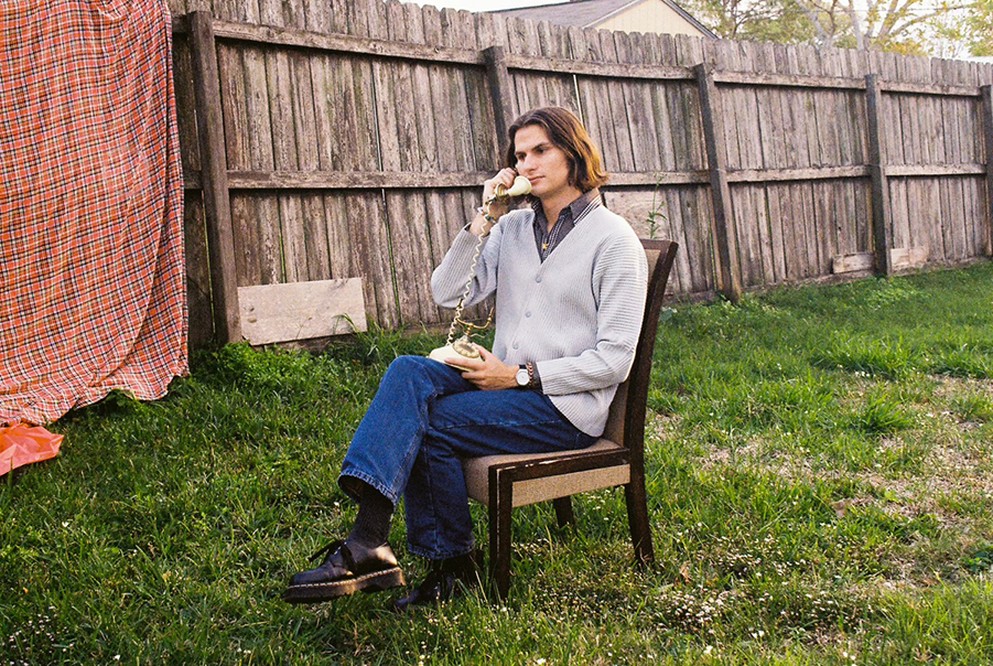 A singer-songwriter sits in a chair in a backyard, holding up an antique-style rotary phone.