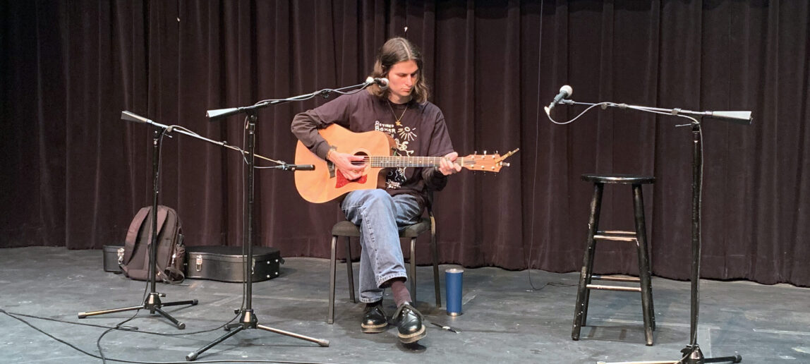 A singer-songwriter performs and plays guitar while seated on a stage.