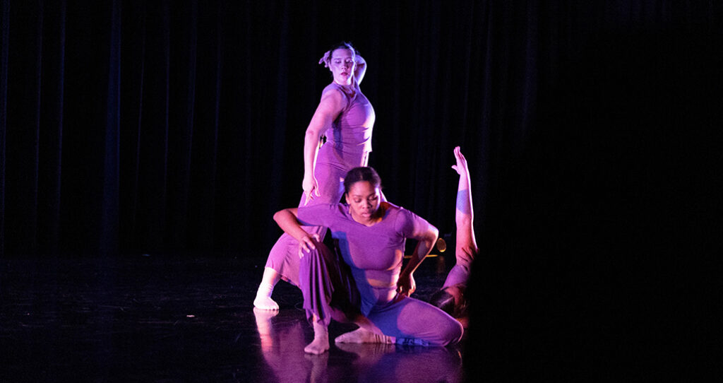 Three dancers perform on a stage. Two are seated, one is standing.