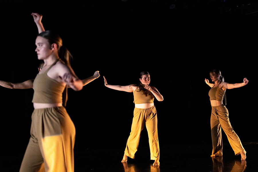 Five dancers in matching brown outfits perform on a stage.