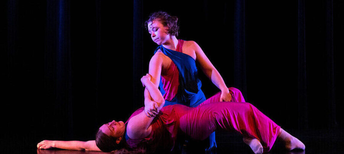 Two dancers perform. One is laying on the floor, while the other has her hands supporting her.