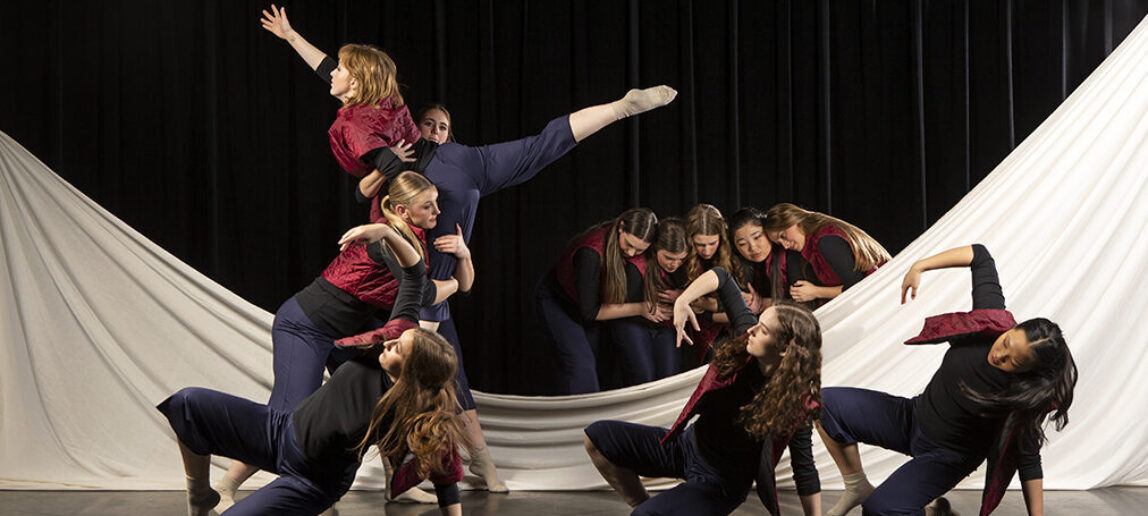 A group of dancers perform on a stage, with a white sheet in the background that has dipped in the middle and is partially on the floor.