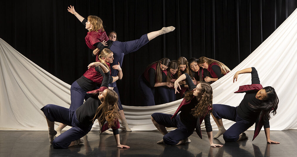 A group of dancers perform on a stage, with a white sheet in the background that has dipped in the middle and is partially on the floor.