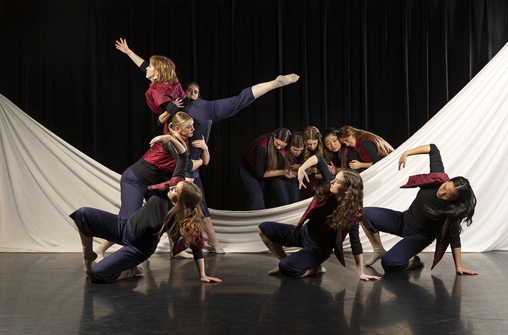 A group of dancers perform on a stage, with a white sheet in the background that has dipped in the middle and is partially on the floor.