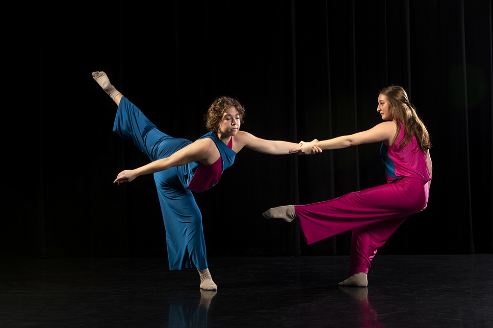 Two dancers, one in blue and one in red. The dancer on the left is in blue, standing on her right foot while her left leg is extended. She is holding the hand of the dancer in red on the right, who is also standing on her right foot.