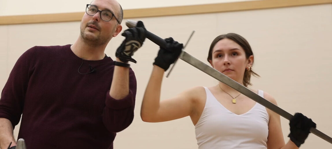 A college student holds a sword in a theatre training exercise, with her professor standing to her right.