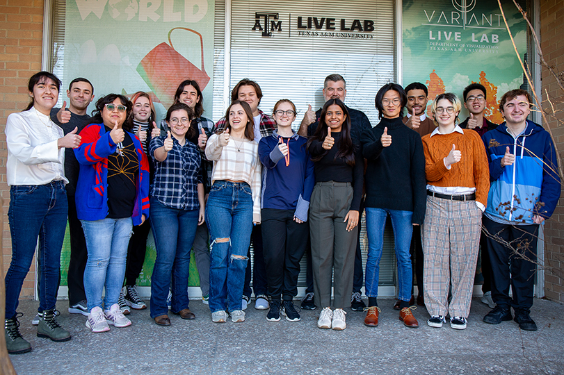 A group of smiling students standing together in front of a window that reads “The LIVE Lab”.